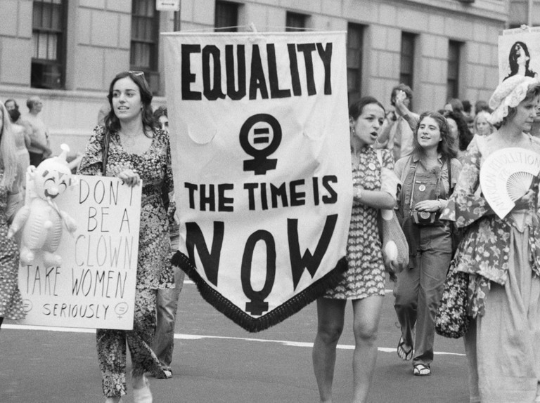 A 1970s women's equality march, carrying a banner reading Equality, The Time Is Now